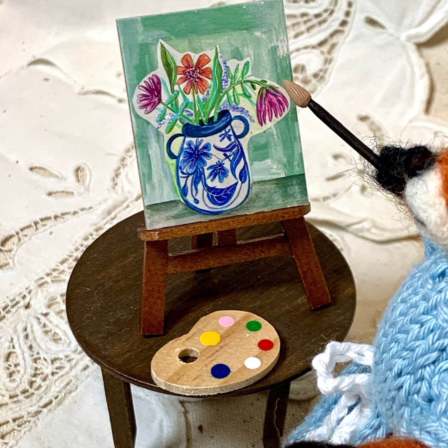 Miniature painting of flowers in a blue vase on an easel with a palette, held by a small creature.