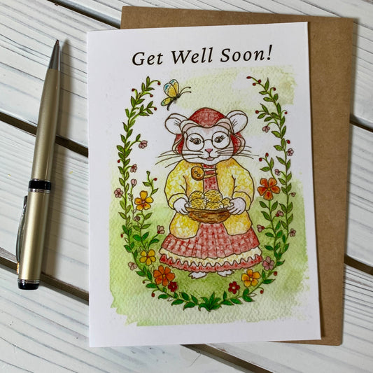 A rectangular greeting card with a watercolor illustration of a mouse in a yellow dress holding a basket of gold cookies, surrounded by flowers and greenery, with the text 'Get Well Soon!' at the top. The card is placed on top of a brown kraft envelope on a white wood surface, with a gold pen beside it.