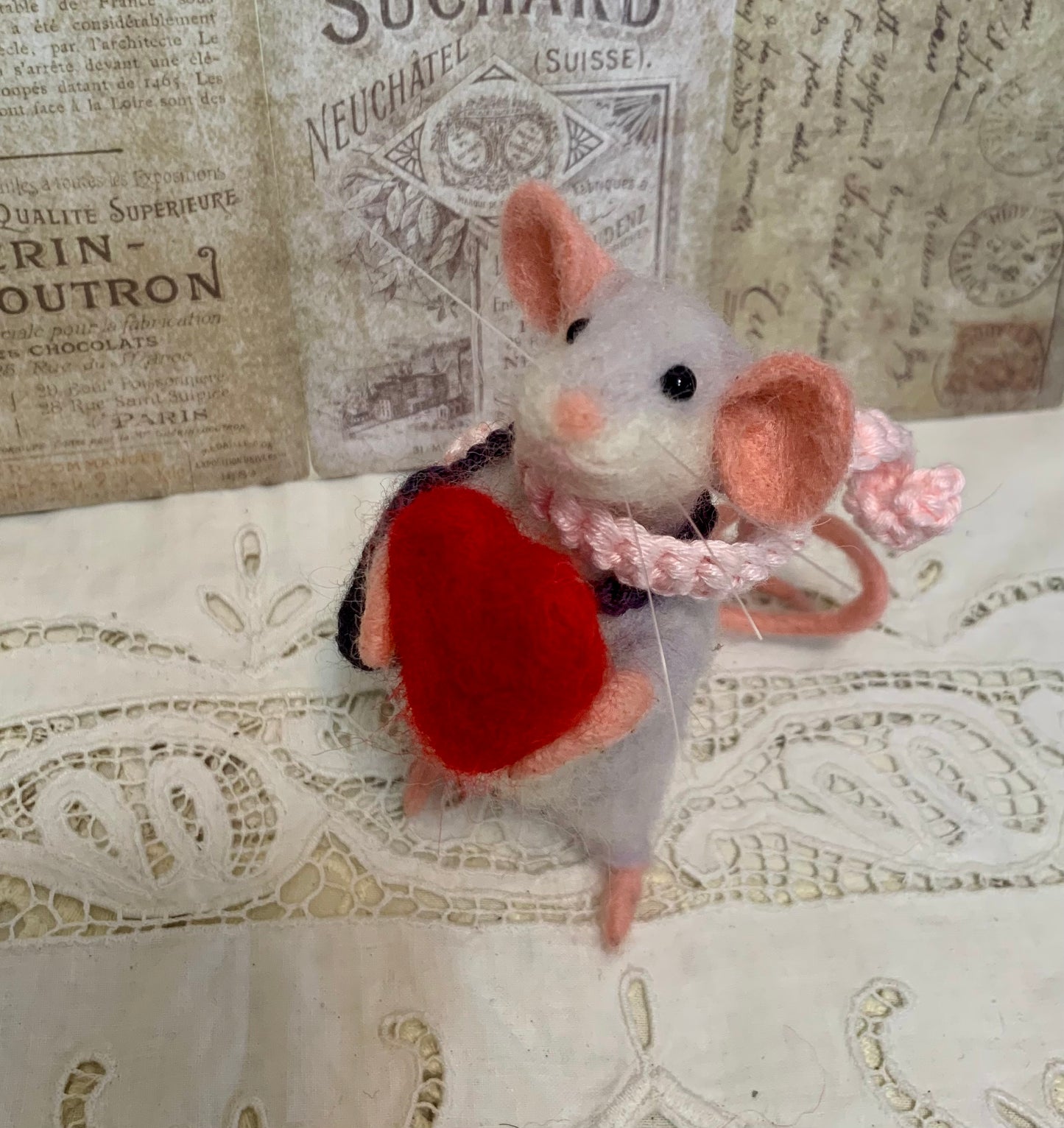 Felted mouse with a red heart and pink bow on a textured surface with vintage newspaper background