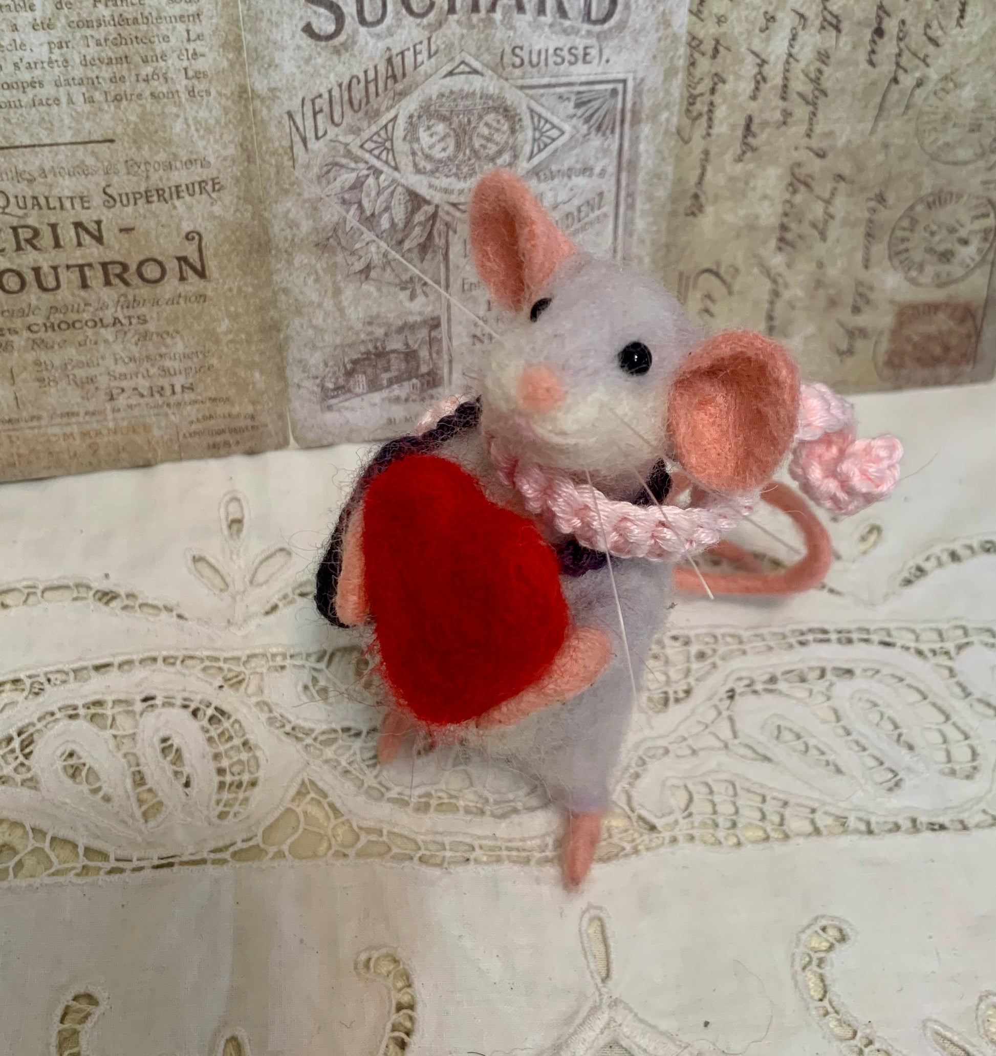Felted mouse with a red heart and pink bow on a textured surface with vintage newspaper background