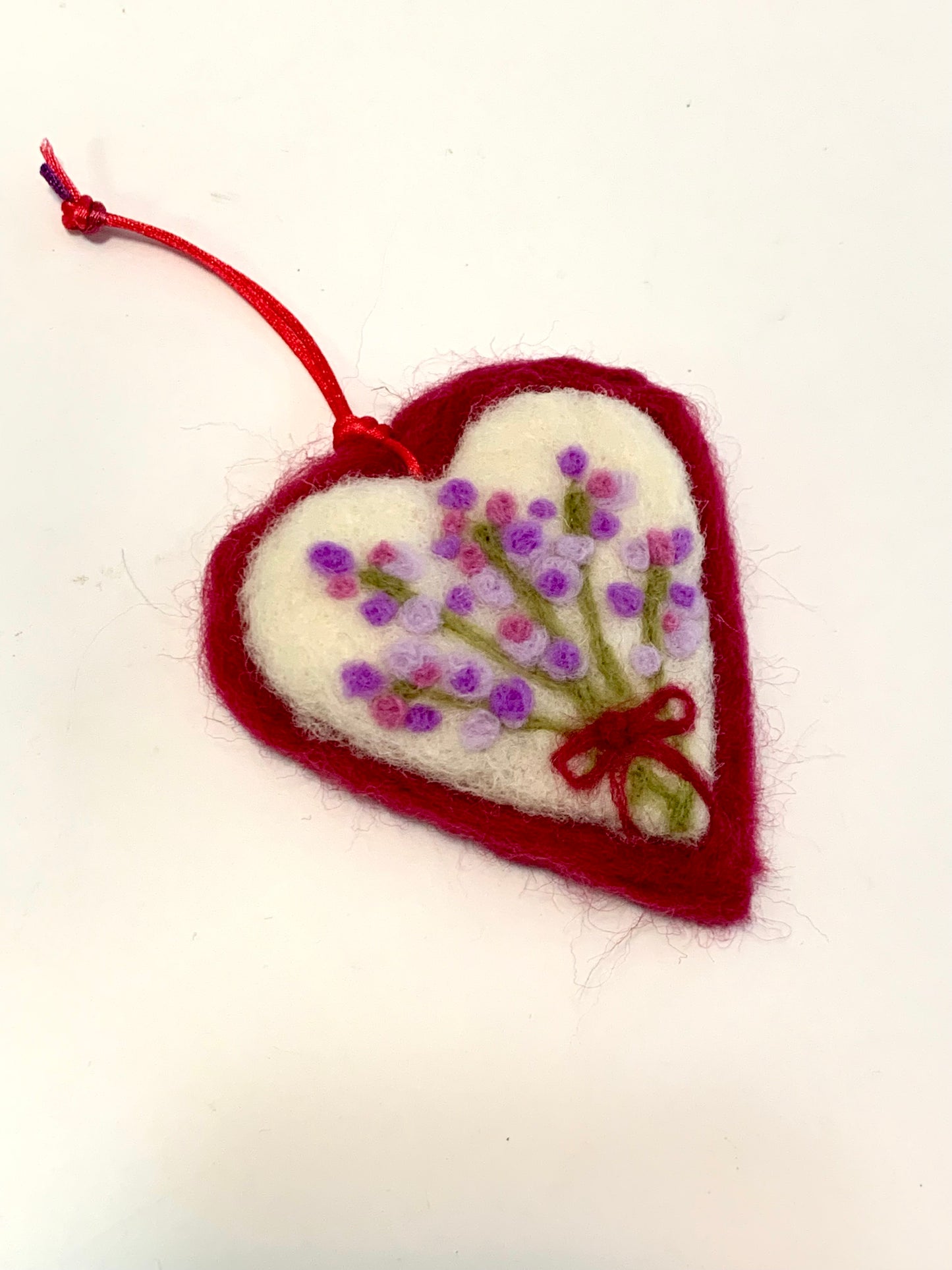 Heart-shaped felt ornament with embroidered flowers and a red ribbon on a white background