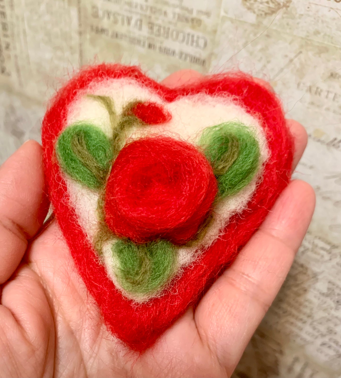 Heart-shaped felted ornament with red rose design held in a hand against a textured background