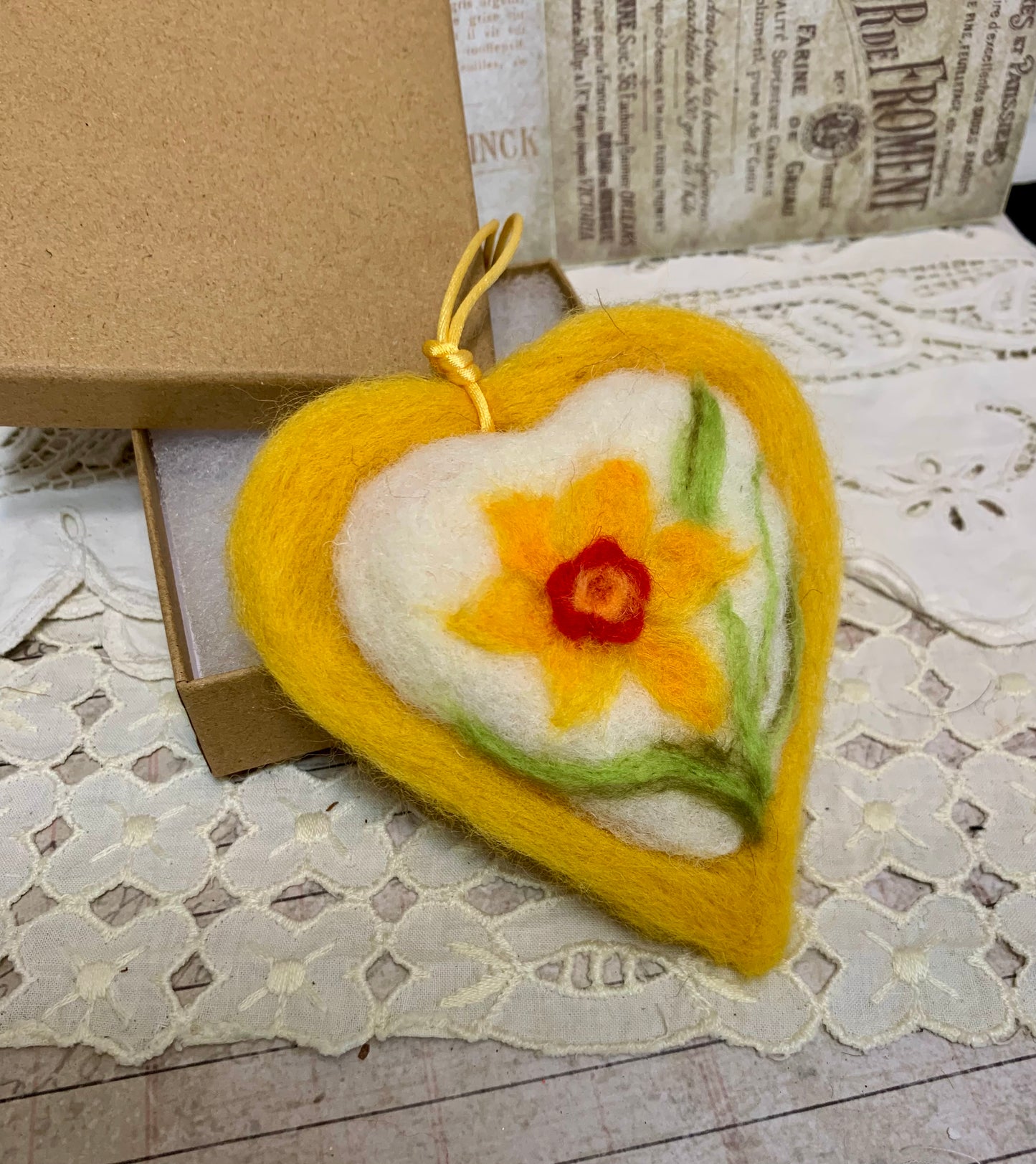 Heart-shaped felted ornament with a daffodil design on a lace surface.