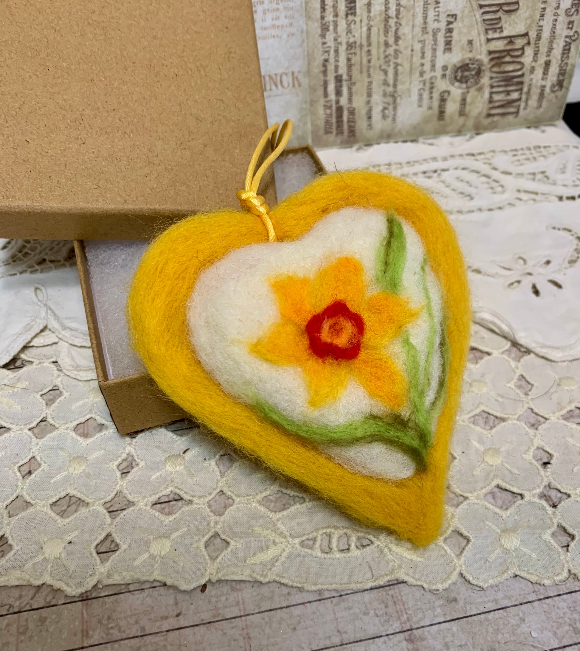 Heart-shaped felted ornament with a daffodil design on a lace surface.