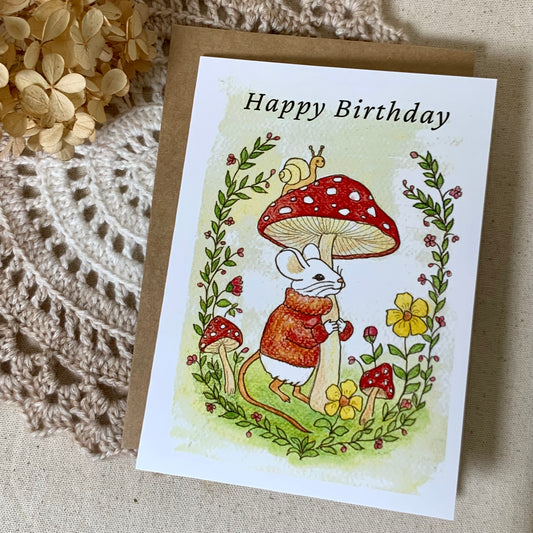 A square birthday card with a brown kraft envelope, featuring a whimsical illustration of a white mouse under a red-and-white mushroom, surrounded by flowers and greenery. The card reads 'Happy Birthday' and is styled on a textured beige surface with a crocheted textile and dried flowers.