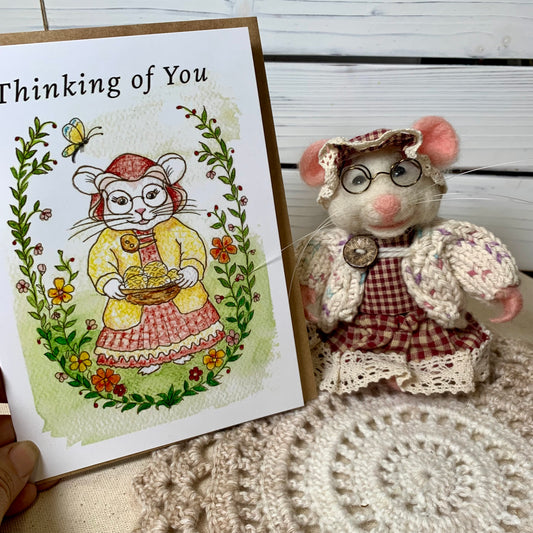 A handmade greeting card with a watercolor illustration of a mouse in a yellow dress holding a basket of honey, next to a matching plush toy mouse dressed in a red gingham dress and lace bonnet. The card reads 'Thinking of You' and is held by a hand over a beige crochet blanket against a white wood paneled background.
