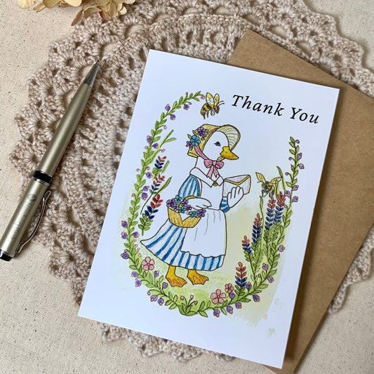 A square white thank-you card with a vintage-style illustration of a duck in a bonnet and dress, surrounded by flowers, placed on a beige textured surface with a brown envelope and a silver pen nearby.