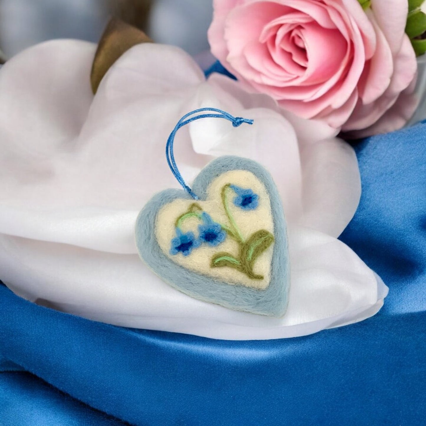 Heart-shaped embroidered ornament with floral design on a white surface with pink roses in the background