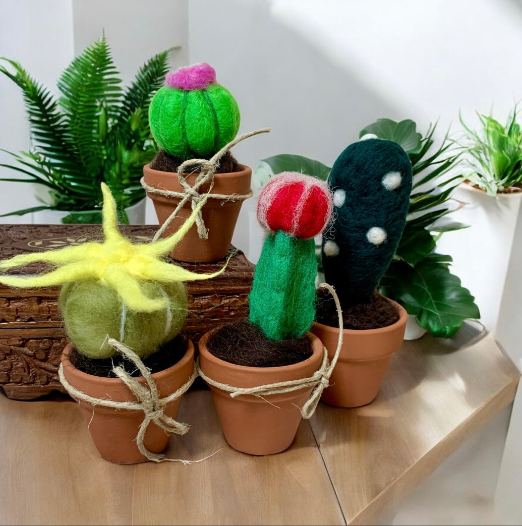 Felted cactus toys in terracotta pots on a wooden surface with plants in the background.