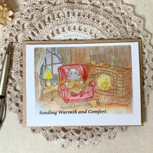 A square greeting card featuring a watercolor illustration of a cat sitting in a red armchair by a fireplace, with the text 'Sending Warmth and Comfort' at the bottom. The card is displayed on a beige fabric surface with a white crocheted doily underneath.