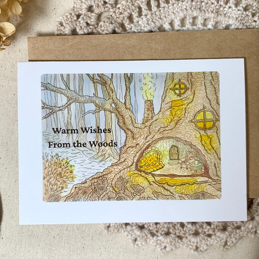 A white greeting card with a brown kraft envelope, featuring an illustration of a tree with small houses in its trunk and the text 'Warm Wishes From the Woods'. The card is laid flat on a textured beige fabric background.