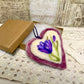 Heart-shaped felted product with a purple flower design on a lace surface with vintage text in the background
