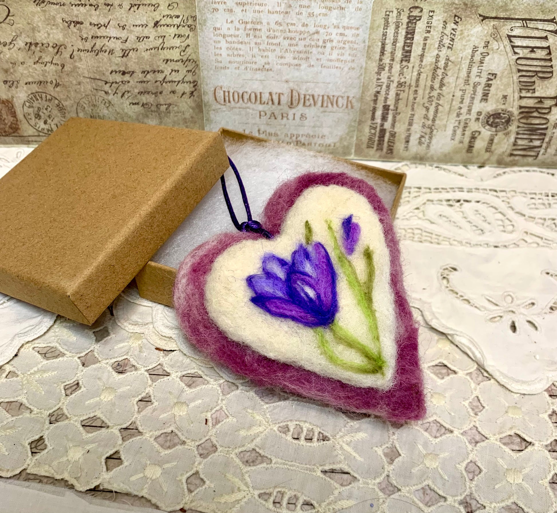 Heart-shaped felted product with a purple flower design on a lace surface with vintage text in the background