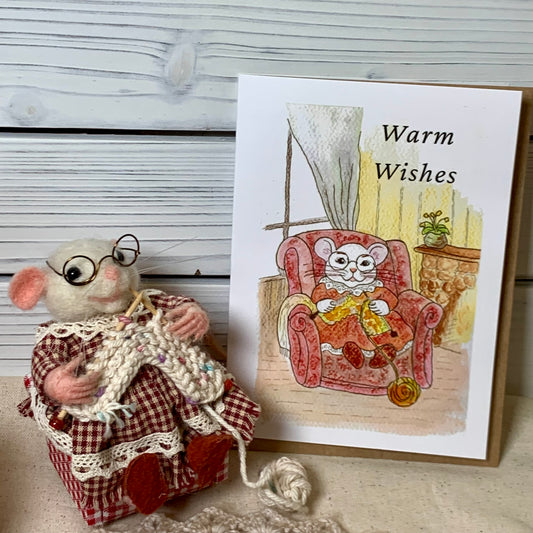 A handmade greeting card featuring a watercolor illustration of a mouse sitting in a red armchair with the text 'Warm Wishes' on the right, next to a plush felt mouse wearing glasses and a red-and-white check knit sweater holding knitting yarn on the left, set against a whitewashed wood background.
