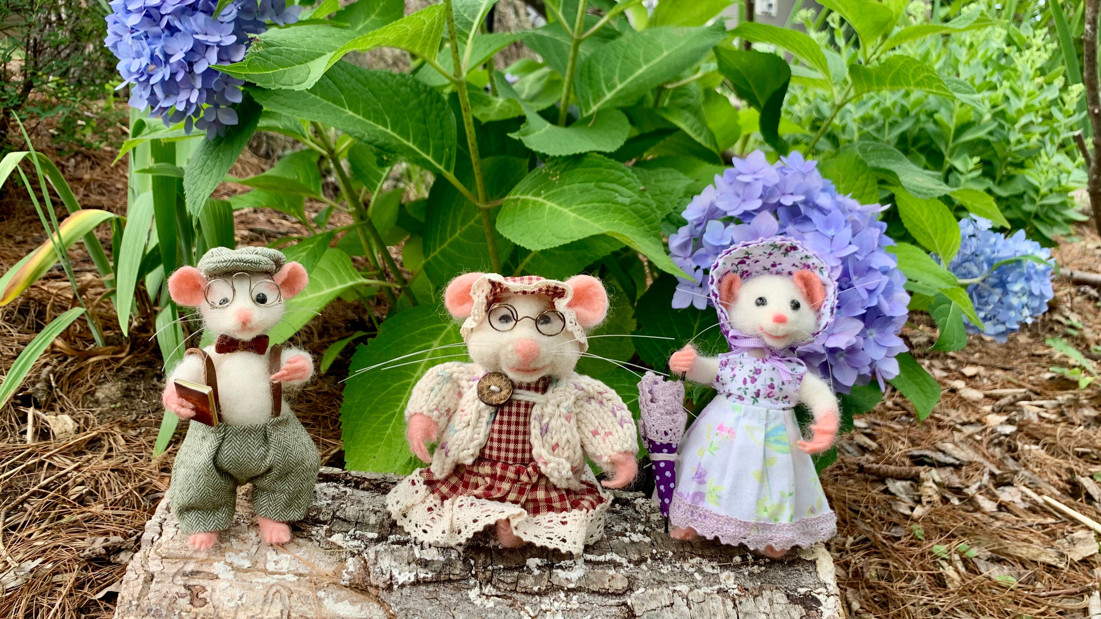 Three small plush mouse figurines dressed in vintage clothing, posed outdoors on a stone surface among green leaves and purple hydrangea flowers.